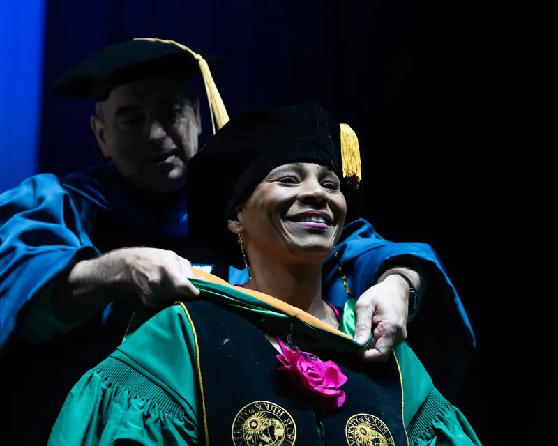 A doctoral graduate in USF green regalia smiles as a faculty member places the doctoral hood over their shoulders at commencement.